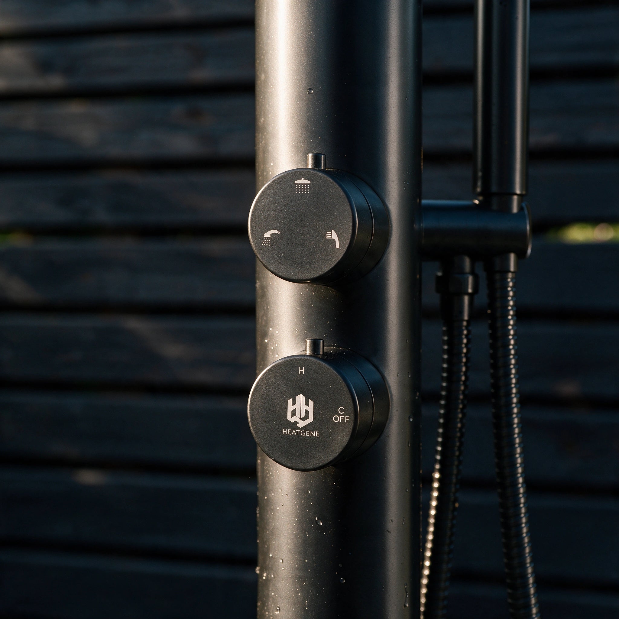 Close-up of a matte black outdoor shower column with two round control dials, one labeled HEATGENE, against a dark horizontal-slat background.
