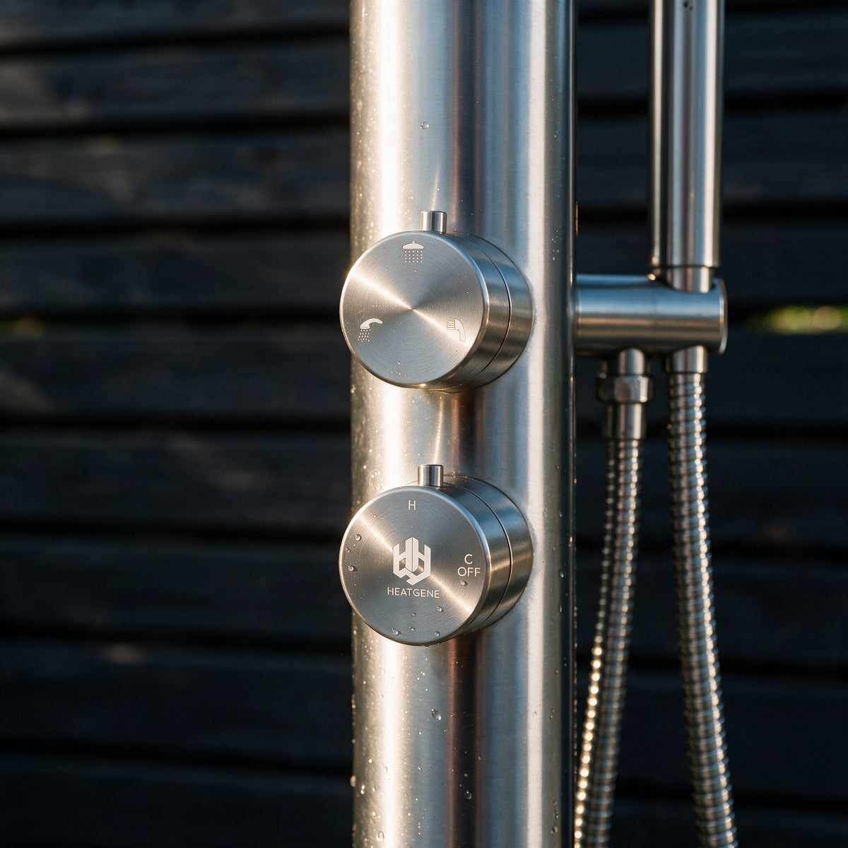 Close-up of a brushed stainless steel outdoor shower column with two round control knobs, one labeled HEATING and one labeled C OFF, against a dark horizontal siding background.