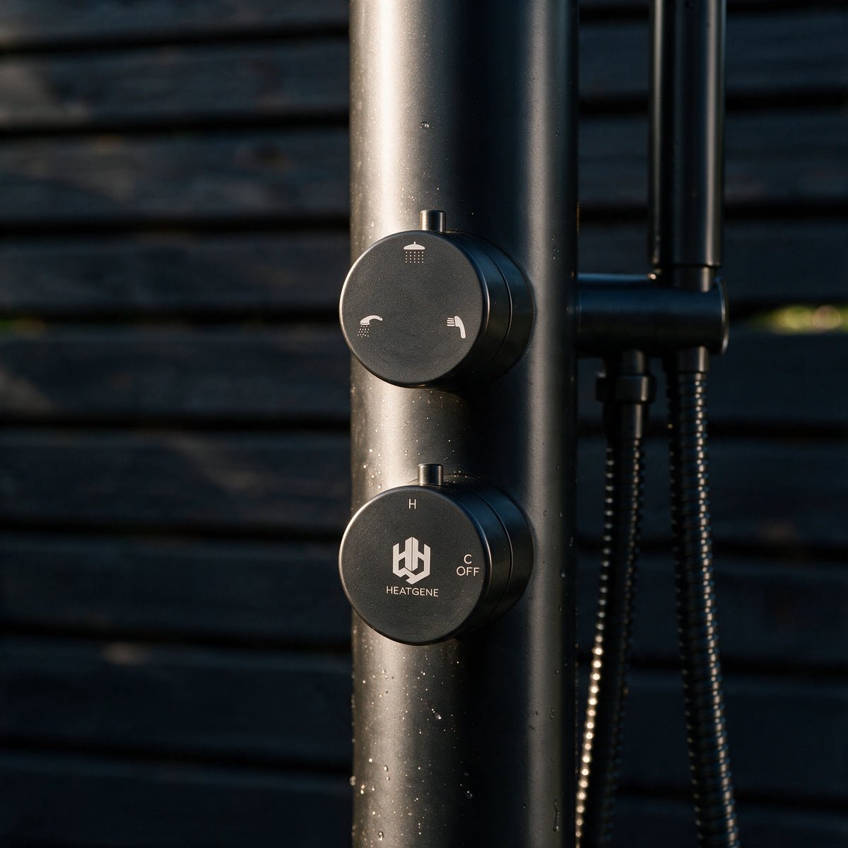 Close-up of a matte black outdoor shower column with two round control knobs, one labeled HEATGENE, set against a dark horizontal slat wall.