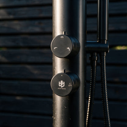 Close-up of a matte black outdoor shower column with two round control knobs, one labeled HEATGENE, set against a dark horizontal slat wall.