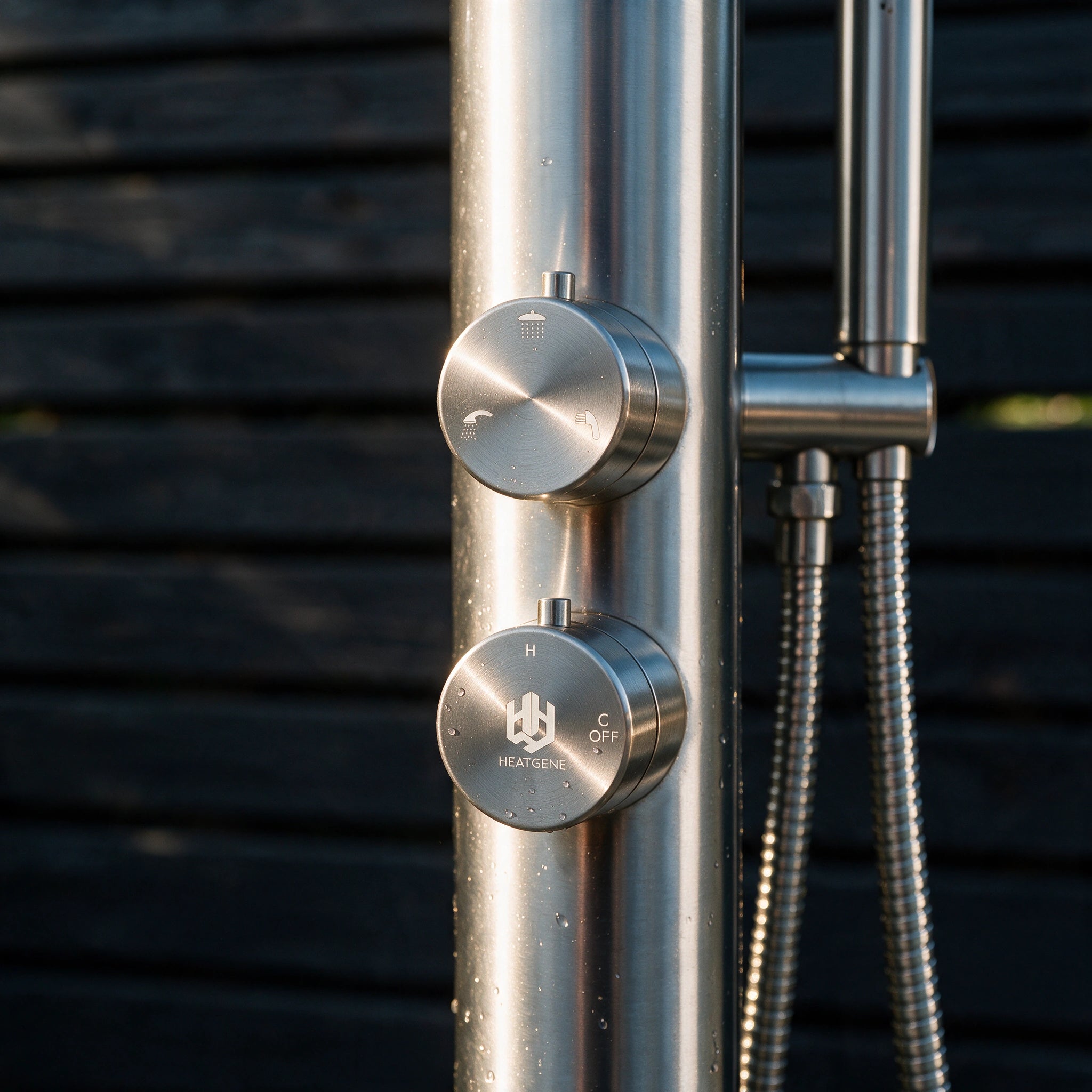 Close-up of a brushed stainless steel outdoor shower column with two round control knobs, one labeled HEATING and one labeled C OFF, against a dark horizontal siding background.