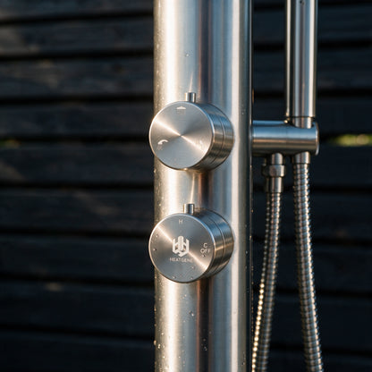 Close-up of a brushed stainless steel outdoor shower column with two round control knobs, one labeled HEATING and one labeled C OFF, against a dark horizontal siding background.